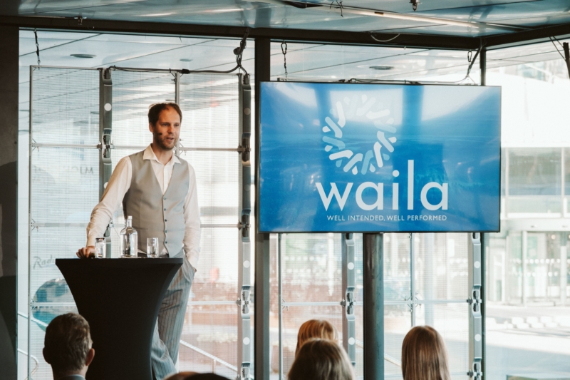 Fotografi av Wailas VD Sebastian Meyer på scen, framför ett podium. Till höger om sig står en TV-skärm med Wailas logotyp. I bakgrunden syns stora fönster ut mot gatan. I bild finns illustrerad symbol föreställande en vindkraft, med texten "Reflektioner från Vind2024"